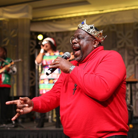 A man in a red shirt and wearing a plastic crown sings into a microphone